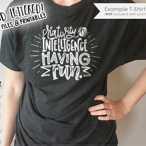 May include: A dark gray t-shirt with a white design that reads "Creativity is intelligence having fun." The design is in a hand-lettered style.