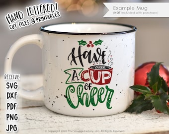 Pode incluir: Uma caneca de cerâmica branca com uma pega preta e um design salpicado. A caneca tem um design verde e vermelho com o texto "Have a Cup of Cheer" em letras pretas e verdes.