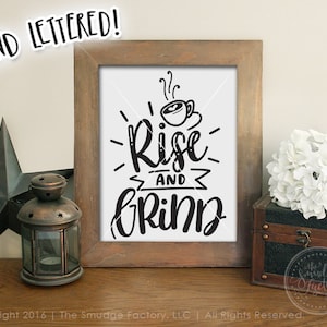 Può includere: Una stampa incorniciata con un design in bianco e nero che dice "Rise and Grind" con una tazza di caffè e vapore che esce da essa. La stampa è in una cornice di legno e si trova su una superficie di legno.