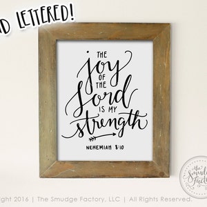 May include: A framed print with a white background and black calligraphy that reads "The joy of the Lord is my strength" with an arrow pointing to the right and the text "Nehemiah 8:10" below.