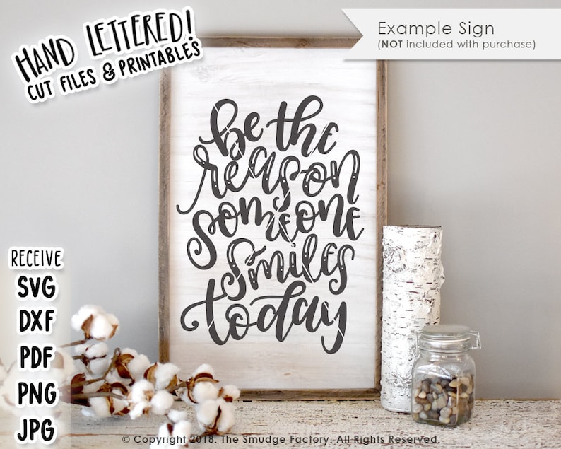 May include: A white wooden sign with black text that reads "Be the reason someone smiles today". The sign is sitting on a white wooden surface with a birch log and a glass jar filled with small rocks.