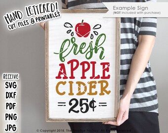 Puede incluir: Un letrero de madera blanco con una manzana roja, hojas verdes y el texto "fresh apple cider = 25¢ =" en estilo de letra manuscrita.