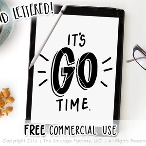 May include: A black tablet displays the phrase "It's Go Time" in bold, hand-lettered style. A white stylus rests on the tablet. The words "Hand Lettered!" and "Free Commercial Use" are also visible. A cup of coffee and goldfish crackers are in the background.