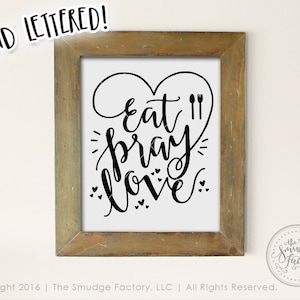 Puede incluir: Una impresión enmarcada con un fondo blanco y caligrafía negra que dice "Eat Pray Love" con una forma de corazón alrededor de las palabras y un gráfico de tenedor y cuchillo. El marco es de madera marrón con un acabado desgastado.