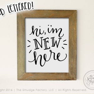 May include: A framed sign with the text "hi, im NEW here" written in black cursive font on a white background. The frame is made of wood with a light brown finish.