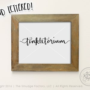 May include: A framed print with the word "Tinkletorium" written in black cursive script. The frame is a light brown wood with a distressed finish. The print is on a white background.