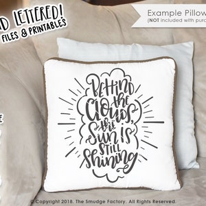 May include: A white pillow with brown trim features a black and white graphic that reads "Behind the clouds the sun is still shining".