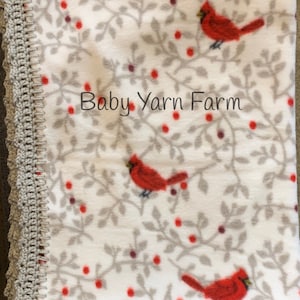 May include: A white fleece baby blanket with a gray crocheted border. The blanket features a repeating pattern of red cardinals and red berries on gray branches. The text "Baby Yarn Farm" is printed in the center of the blanket.