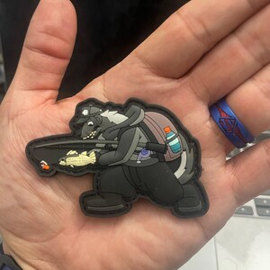 Pode incluir: Um patch de desenho animado preto e branco de um texugo usando um chapéu e segurando uma vara de pesca. O texugo está usando uma mochila e tem uma garrafa de água presa a ela. O patch está na mão de uma pessoa.