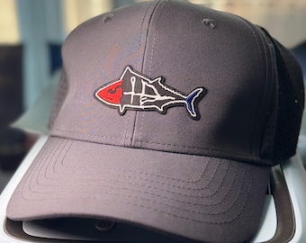 May include: A grey baseball cap featuring an embroidered patch of a stylized fish on the front. The fish design is outlined in black and white, with a red head section and a blue tail tip. The cap has a structured crown and a curved brim.