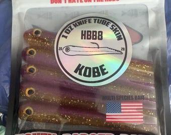 May include: A clear plastic package containing five purple and gold glitter fishing lures. The label reads DON'T HATE ON THE HBB8, 1 OZ KNIFE TUBE SKIN, HBB8, KOBE, MULTI SPECIES BAIT, and HONEY BADGER BAITS, with a small American flag graphic.