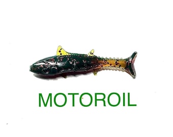 May include: A soft plastic fishing lure shaped like a small fish with a dark green body, yellow fins, and a translucent tail section containing red glitter flecks. The word MOTOROIL is printed in green capital letters below the lure.