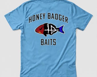 May include: A light blue t-shirt featuring a graphic of a fish with a red head and blue tail. The text HONEY BADGER is printed in black above the fish, and the word BAITS is printed in black below the fish graphic on the back of the shirt.