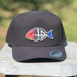 Puede incluir: Una gorra snapback gris carbón con un gráfico de pez estilizado en el panel frontal. El diseño del pez está segmentado con una cabeza roja, una sección media esquelética blanca y una cola azul. Una pegatina circular en la visera dice SNAPBACK.