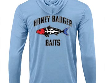 May include: A light blue long-sleeve hooded shirt featuring a graphic of a fish with a red head and blue tail. The text HONEY BADGER is printed in black above the fish, and the word BAITS is printed in black below the fish graphic.
