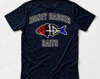 May include: A navy blue short-sleeve t-shirt featuring the white text HONEY BADGER BAITS. Centered is a graphic of a fish silhouette with a red head, a white skeletal midsection, and a blue tail fin, all outlined in white on the back.