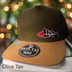 May include: An olive green baseball cap with a tan brim and a fish-shaped patch featuring red and white details. A circular black sticker on the brim displays the text BASIC CAP 5010 HEADWEAR SUPPLY LOS ANGELES CALIFORNIA USA.
