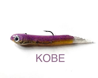 May include: A purple and gold fishing lure with a glitter finish, a silver eye, and a black hook. The lure features a translucent tail and the text KOBE printed in purple below the body on a plain white background.