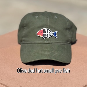 Op de afbeelding: Een olijfgroene baseballpet met een kleine PVC-patch aan de voorzijde. De patch toont een gestileerde visafbeelding verdeeld in secties van rood, zwart en blauw met witte omlijningen. De tekst Olive dad hat small pvc fish is zichtbaar.
