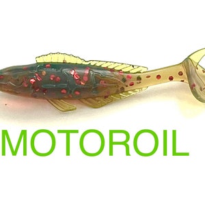 May include: A green and brown plastic fishing lure with red glitter. The lure is shaped like a fish and has a tail that is bent at a 90-degree angle. The lure is labeled "MOTOROIL".