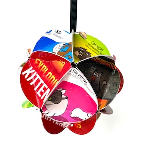 Peut inclure: Une décoration de jeu de cartes Exploding Kittens rouge, verte, bleue, noire et rose. La décoration est faite de cartes pliées du jeu et comporte le texte "Exploding Kitten" et "Om Nom".