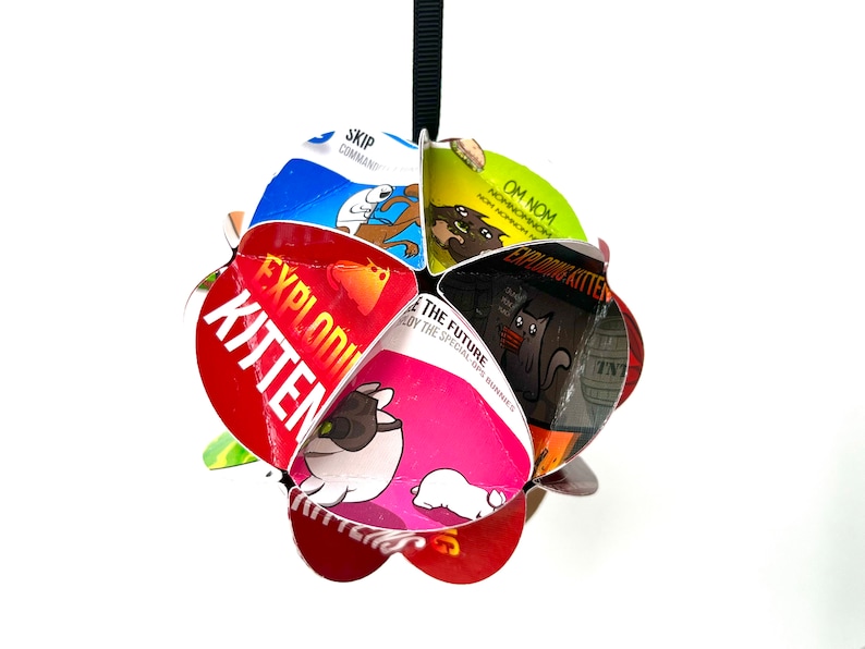May include: A red, green, blue, black, and pink Exploding Kittens card game ornament. The ornament is made of folded cards from the game and features the text "Exploding Kitten" and "Om Nom".