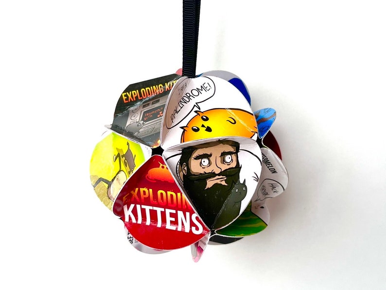 May include: A multi-colored paper ornament shaped like a flower with the words "Exploding Kittens" printed on it. The ornament features a cartoon image of a bearded man with a cat in his beard.