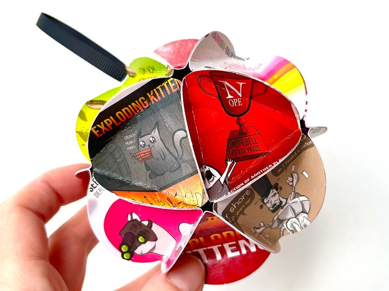 May include: A red, white, and black Exploding Kittens card game ornament with a black cat holding a stick of dynamite. The ornament is shaped like a star and has a black string attached to it.