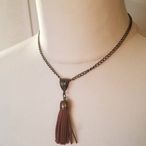 May include: A bronze-toned necklace with a chain and a pendant featuring a brown suede tassel. The tassel is attached to a decorative metal cap. The necklace is displayed on a mannequin.