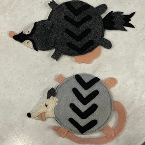 May include: Two felt animal appliques, a raccoon and a possum, with black chevron patterns on their backs. The raccoon is gray with a black nose and eyes, and the possum is gray with a white nose and eyes. Both animals have pink felt tails.