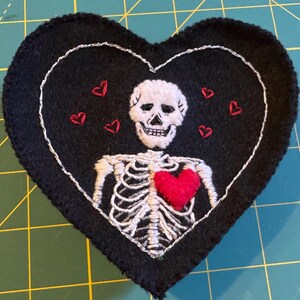 May include: A black felt heart-shaped patch with a white embroidered skeleton holding a red felt heart. The skeleton has a skull with a white embroidered smile. There are five small red embroidered hearts around the skeleton.