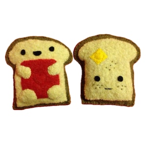 Może przedstawiać: Dwie pluszowe kromki chleba, jedna z czerwonym dżemem, a druga z żółtym masłem. Kromki chleba mają wyszywane oczy i uśmiechy.