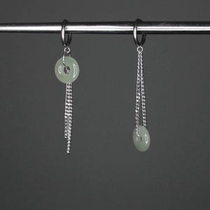 May include: A pair of silver earrings with a light green jade stone charm on each. The charms are round and have a hole in the center. The earrings are hanging from a silver chain.