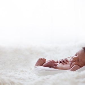 Peut inclure: Un nouveau-né aux cheveux foncés dort paisiblement sur une surface douce, blanche et duveteuse. Le bébé est enveloppé dans un tissu blanc, seules la tête et les pieds sont visibles. L'image est éclairée doucement.