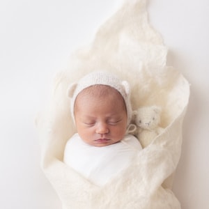 Puede incluir: Un bebé recién nacido que lleva un gorro blanco de punto con orejeras está durmiendo plácidamente en un arrullo blanco. Un pequeño animal de peluche blanco está acurrucado junto al bebé.