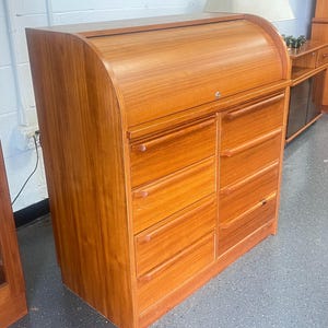 May include: A medium-brown wooden roll-top desk featuring a curved sliding tambour door at the top and six stacked drawers below. The drawers are arranged in two vertical columns of three, each equipped with horizontal wooden pull handles.