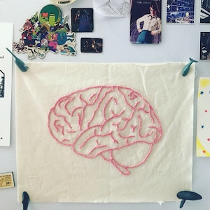 May include: A pink embroidered brain on a white fabric background. The brain is stitched with pink thread and has a detailed design.