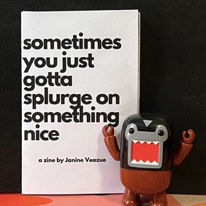 May include: A small, white zine with the text "sometimes you just gotta splurge on something nice" and the text "a zine by Janine Veazue" at the bottom. A brown and black Domo figure stands next to the zine.