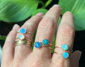 May include: A collection of gold rings, each featuring a round opal stone. The opals vary in color, showcasing shades of blue and white. The rings are displayed on a hand, with a green leafy background.