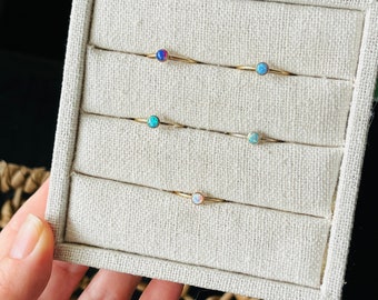 May include: A collection of delicate gold rings, each featuring a small, round opal stone in various colors. The opals range from blue and green to pink and white. The rings are displayed in a beige jewelry tray.