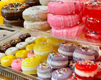 Puede incluir: Una vitrina llena de jabones decorativos coloridos con forma de donuts y macarons. Los donuts tienen varios toppings, incluyendo chispas y glaseado. Los macarons son amarillos, rosas y morados con relleno blanco y chispas. Los jabones están dispuestos en estantes transparentes.