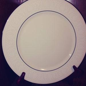 May include: A white ceramic dinner plate with a delicate floral pattern around the rim. The plate has a black rim and is on a wooden stand.