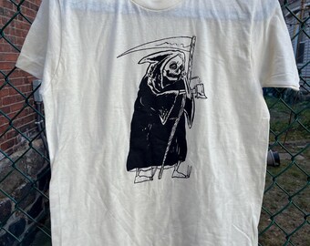 May include: Off-white t-shirt featuring a black graphic of the Grim Reaper holding a scythe and a bell. The illustration is detailed, with the Reaper's skeletal face and hooded cloak. The shirt is hanging on a black hanger.