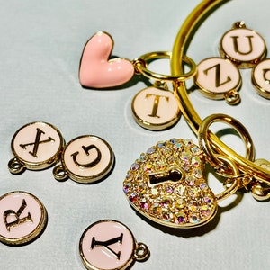 May include: A gold-colored bracelet with a heart-shaped charm and several letter charms. The heart charm is encrusted with iridescent crystals. Additional charms include the letters A, G, R, T, U, X, Y, and Z. The charms have a pink background and gold trim.
