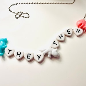 May include: A necklace with a silver chain and a series of white heart-shaped beads spelling out "THEY THEM". The necklace features a blue and pink dinosaur charm on each end and a white dinosaur charm in the middle.