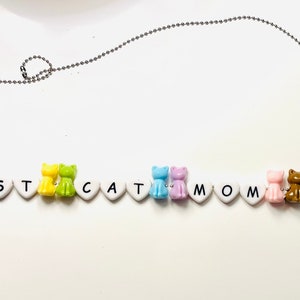 May include: A beaded necklace with the phrase "BEST CAT MOM EVER" spelled out with white heart-shaped beads. Colorful cat-shaped beads in pink, orange, yellow, green, blue, purple, and gold accent the necklace.