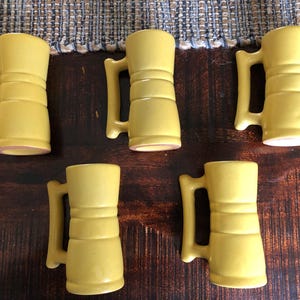 May include: Five yellow ceramic mugs with handles are arranged on a dark wooden surface. The mugs have a cylindrical shape with horizontal ridges and a handle on the side. The mugs are a light yellow colour.