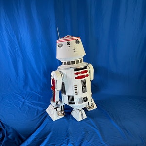 May include: A white and red robot with a dome-shaped head and a silver body. The robot has two red stripes on its chest and is standing on a blue background.