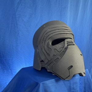 Puede incluir: Un casco de plástico negro con un diseño detallado, que se asemeja a un personaje de una película de ciencia ficción. El casco está sentado sobre una superficie de tela azul.