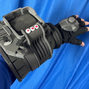 May include: A black and gray foam prop resembling a futuristic arm brace with a small gray device on the hand. The brace has a series of black ridges and a small black knob on the side. There are three red circles on the top of the brace.